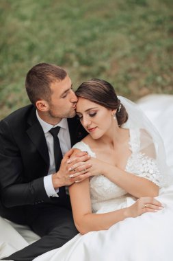 Gelin ve damat battaniyenin üzerinde oturuyor, gelin beyaz bir elbise giyiyor ve damat siyah bir takım elbise giyiyor.