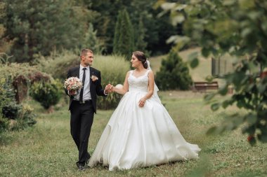 Bir gelin ve damat parkta yürüyorlar. Gelin beyaz bir elbise giyiyor ve damat bir takım elbise giyiyor. El ele tutuşuyorlar ve mutlu görünüyorlar.