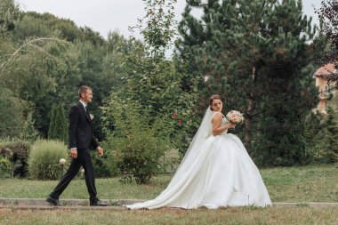 Bir gelin ve damat parkta bir patikada yürüyorlar. Gelin beyaz bir elbise giyiyor ve elinde bir buket tutuyor. Sahne huzurlu ve romantik.