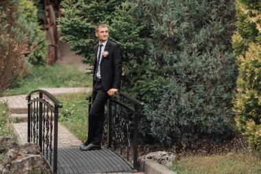 Takım elbiseli bir adam köprüde duruyor. Bir kadınla el ele tutuşuyor. Sahne romantik ve samimi.