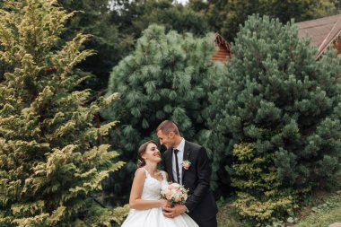 Bir gelin ve damat ağaçlı bir ormanın önünde duruyorlar. Gelin bir buket tutuyor ve damat kravat takıyor. Sahne mutlu ve romantik.