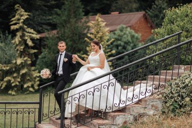 Bir gelin ve damat merdivenlerden iniyorlar. Gelin beyaz bir elbise giyiyor ve damat bir takım elbise giyiyor. El ele tutuşuyorlar ve mutlu görünüyorlar.