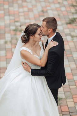 Bir gelin ve damat tuğla kaldırımda öpüşüyorlar. Gelin beyaz bir elbise giyiyor ve damat siyah bir takım elbise giyiyor. Sahne romantik ve mutlu.