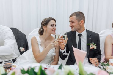 Bir gelin ve damat düğün resepsiyonunda kadeh kaldırıyor. Gelin beyaz bir elbise giyiyor ve damat siyah bir takım elbise giyiyor. İkisi de ellerinde şarap kadehleriyle kameraya gülümsüyor.