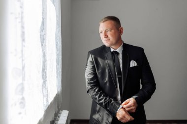 Takım elbiseli bir adam pencerenin önünde duruyor, sokağa bakıyor. O bir kravat ve siyah bir ceket giyiyor.