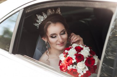 Bir gelin evlenmeye hazırlanıyor ve bir arabada oturuyor. O bir taç takıyor ve kucağında bir buket kırmızı gül var.