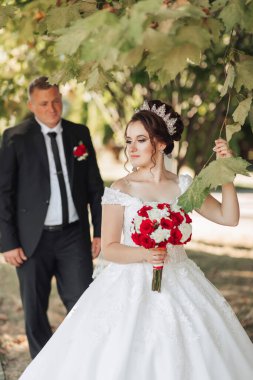 Bir gelin ve damat parkta duruyorlar, gelin elinde bir buket kırmızı çiçek tutuyor.
