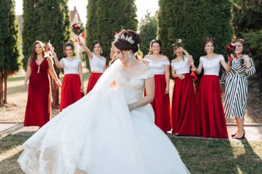 Bir gelin ve nedimeleri parkta fotoğraf çektirmek için poz veriyorlar. Gelin beyaz bir elbise ve taç giyerken nedimeler kırmızı elbiseler giyiyor.