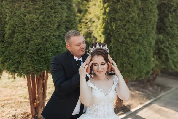 Bir gelin ve damat bir ağacın önünde fotoğraf için poz veriyorlar. Gelin taç takıyor ve damat da takmasına yardım ediyor. Sahne mutlu ve romantik.