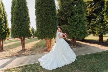 Beyaz elbiseli bir kadın bir sıra ağacın önünde duruyor. Elinde bir buket kırmızı çiçek tutuyor.