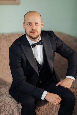 Takım elbiseli ve kravatlı bir adam kanepede oturuyor. Siyah bir takım elbise ve siyah papyonlu beyaz bir gömlek giyiyor.