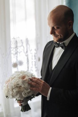 Siyah takım elbiseli bir adam elinde bir buket beyaz çiçek tutuyor. Adam gülümsüyor ve mutlu.