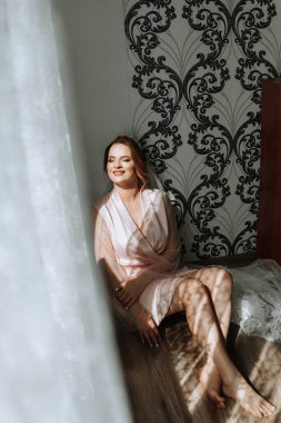 Pembe elbiseli bir kadın arkasında penceresi olan bir yatakta oturuyor. Gülümsüyor ve gün ışığının tadını çıkarıyor.