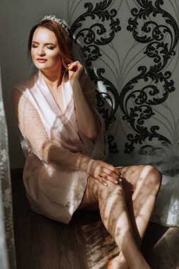 Pembe elbiseli bir kadın elleri dizlerinin üzerinde yerde oturuyor. Bir taç ve küpe takıyor.