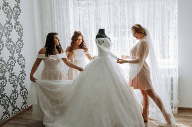 Üç kadın düğün için hazırlanıyor. Bir tanesi elbise tutuyor ve diğer ikisi de ona yardım ediyor.