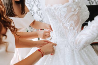 Üç kadın düğün için hazırlanıyor. Bir tanesi gelinin elbisesini ayarlıyor. Diğer iki kadın arkasında durup ona yardım ediyor.