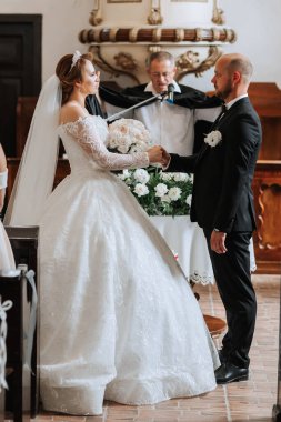 Bir gelin ve damat elinde İncil tutan bir rahibin önünde duruyor. Gelin beyaz bir elbise giyiyor ve damat siyah bir takım elbise giyiyor. Sahne bir düğün töreni.