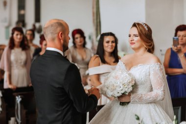 Bir gelin ve damat kilisede evleniyor. Gelin beyaz bir elbise giyiyor ve damat bir takım elbise giyiyor. Gelin bir buket çiçek tutuyor ve damat elini tutuyor.