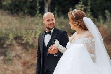 Bir adam ve bir kadın tarlada dikiliyorlar, adam siyah takım elbise giyiyor ve kadın beyaz bir elbise giyiyor.