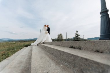 Bir gelin ve damat basamaklarda öpüşüyorlar. Gelin beyaz bir elbise giyiyor ve damat siyah bir takım elbise giyiyor. Sahne arka planda bulutlu bir gökyüzü olan bir alanda geçiyor.