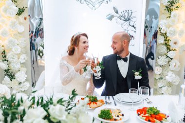 Bir gelin ve damat düğünlerini kadeh kaldırarak kutluyorlar. Masa, şarap bardakları ve bir şişe de dahil olmak üzere çeşitli yiyecek ve içeceklerle hazırlandı. Çift çiçekler ve bir aynayla çevrili.