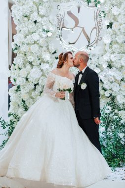 Bir gelin ve damat çiçeklerle beyaz bir duvarın önünde öpüşüyor. Gelin uzun beyaz bir elbise giyiyor ve damat siyah bir takım giyiyor.