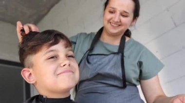 Bir kadın bir çocuğun saçını kesiyor. Çocuk gülümsüyor. Kadın siyah bir önlük giyiyor.