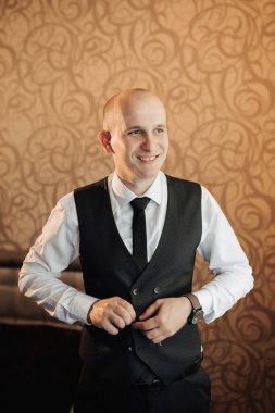 Siyah yelek ve beyaz gömlekli bir adam gülümsüyor ve kravatını düzeltiyor. Adam takım elbise ve kravat giydiği için güven ve profesyonellik kavramı. Adamın gülümsemesi mutlu olduğunu gösteriyor.