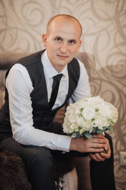 Takım elbiseli ve kravatlı bir adam elinde bir buket beyaz çiçekle koltukta oturuyor.