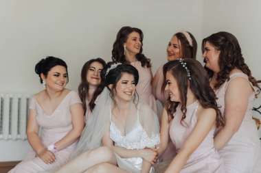 Bir grup kadın birlikte oturuyor, içlerinden biri gelinlik giyiyor. Hepsi gülümsüyor ve birbirlerine eşlik etmekten zevk alıyor gibi görünüyorlar.