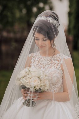 Bir gelin peçe takıyor ve elinde bir buket beyaz çiçek tutuyor. Gülümsüyor ve kameraya bakıyor.