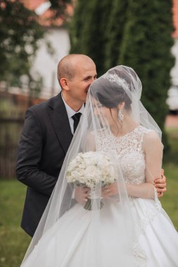 Bir gelin ve damat birbirlerine sarılıp öpüşüyorlar. Gelin bir duvak ve beyaz bir elbise giyiyor.