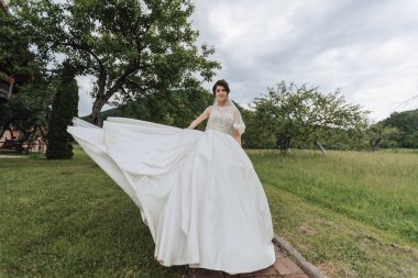Beyaz elbiseli bir gelin, arkasında bir ağaçla bir tarlada duruyor. Elbisesini kaldırıyor ve kameraya bakıyor. Sahne neşeli ve kutlayıcıdır.