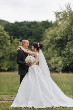 Gelin ve damat tarlada duruyorlar, gelin beyaz bir elbise giyiyor ve damat takım elbise giyiyor.