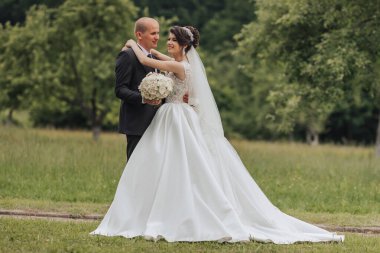 Gelin ve damat tarlada duruyorlar, gelin beyaz bir elbise giyiyor ve damat da takım elbise giyiyor.