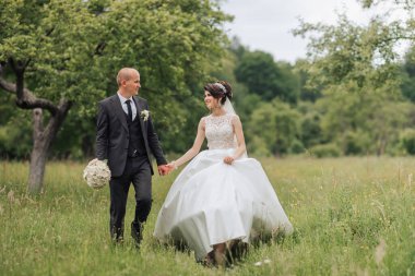 Bir gelin ve damat çim tarlasında yürüyor. Gelin beyaz bir elbise giyiyor ve damat bir takım elbise giyiyor. El ele tutuşuyorlar ve mutlu ve aşık görünüyorlar.