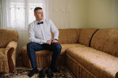 Takım elbiseli ve papyonlu bir adam kanepede oturur. O bir saat takıyor ve elleri kucağında. Kanepe kahverengi ve çiçek desenli.