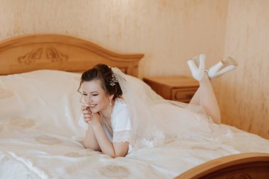 Bir gelin elleri birbirine kenetlenmiş bir şekilde yatakta yatıyor. Gülümsüyor ve mutlu.