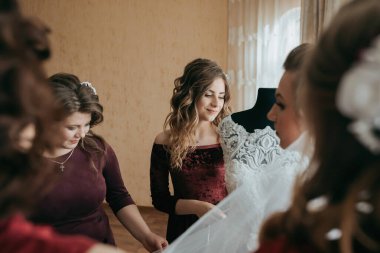 Bir grup kadın bir gelinliğin etrafında toplanıyor ve bir tanesi duvak tutuyor. Sahne bir kutlama ve heyecandır.