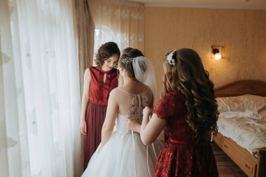 Üç kadın düğün için hazırlanıyor. Biri beyaz bir elbise giyiyor ve diğer ikisi kırmızı elbiseler giyiyor. Bir pencerenin önünde duruyorlar, gelinin elbisesini düzeltiyorlar.