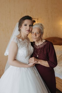 Gelinlik giymiş genç bir kadın yaşlı bir kadının yanında duruyor. Sahne sıcak ve sevgi dolu, çünkü iki kadın aynı anı paylaşıyor.