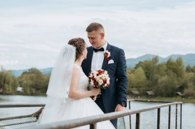 Bir gelin ve damat, bir su kütlesine tepeden bakan bir iskelede duruyor. Gelin bir buket çiçek tutuyor ve damat bir takım elbise giyiyor. Sahne romantik ve resimli.
