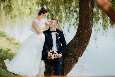 Bir gelin ve damat bir su kütlesinin altında oturuyor. Gelin bir buket tutuyor ve damat kravat takıyor. Sahne romantik ve huzurlu.