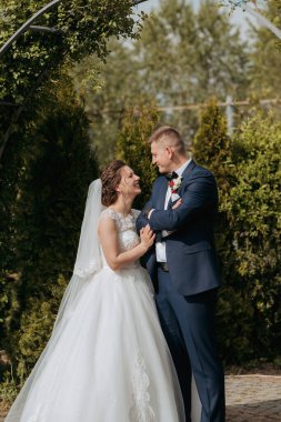 Bir gelin ve damat bir çalılığın önünde kameraya gülümsüyor. Gelin beyaz bir elbise giyiyor ve damat mavi bir takım elbise giyiyor. Sahne mutlu ve neşeli.