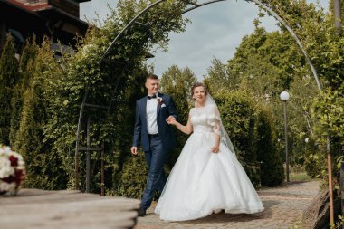 Bir gelin ve damat bahçede bir patikada yürüyorlar. Gelin beyaz bir elbise giyiyor ve damat da takım elbise giyiyor.