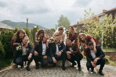 Bir gelin ve damat da dahil olmak üzere bir grup insan resim için poz veriyor. Gelin kırmızı bir elbise giyiyor ve damat bir takım elbise giyiyor. Grup tuğla kaldırımda duruyor.