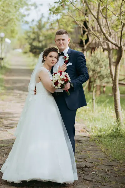 Bir gelin ve damat bir ağacın önünde fotoğraf için poz veriyorlar. Gelin beyaz bir elbise giyiyor ve damat mavi bir takım elbise giyiyor. Bir buket ve kırmızı bir gül tutuyorlar.