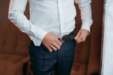 Bir adam beyaz gömlek ve mavi pantolon giyiyor. O, kemerini ayarlıyor. Formalite kavramı ve detaylara dikkat