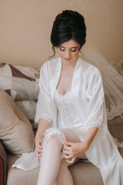 Beyaz önlüklü bir kadın kanepeye oturup çoraplarını düzeltiyor. Kadın gelinlik giydiği için zarafet ve resmiyet kavramı.