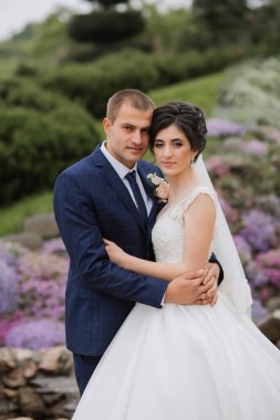 Bir gelin ve damat mor çiçek tarlasının önünde poz veriyorlar. Gelin beyaz bir elbise giyiyor ve damat mavi bir takım elbise giyiyor. Sahne mutlu ve romantik.